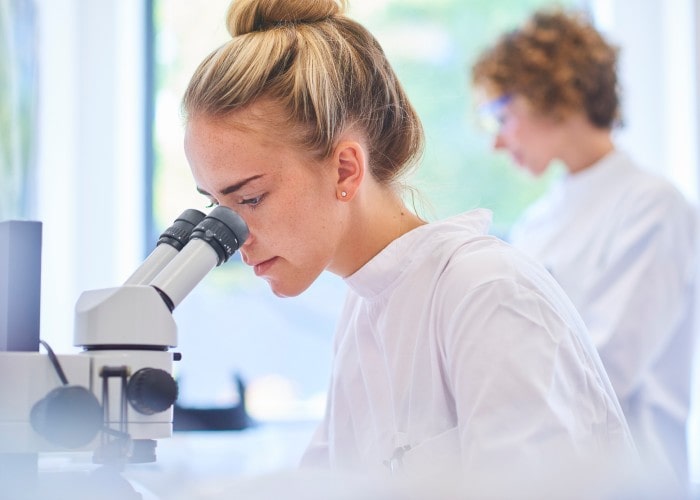 Kvinne i hvitt labutstyr ser ned i mikroskop